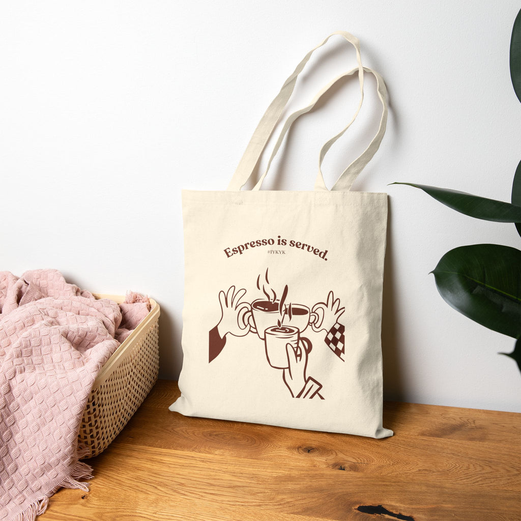 Canvas tote bag featuring an Espresso is served graphic with three clinking coffee mugs on the back and a front pocket W logo from The W-League collection
